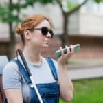 Woman wearing sunglasses and denim overalls sits on a park bench outdoors, talking on a mint green smartphone with a white cane resting at her side.