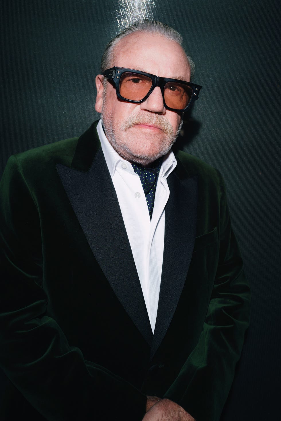 ray winstone wearing sunglasses and a velvet jacket, standing against a dark background.