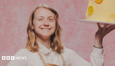 Bake-Off promo studio shot of Danni standing in a pink studio holding up a yellow iced cake on a board with one hand. They have shoulder length dark blonde hair and are wearing a long sleeved blouse with a lace collar and a beige apron.