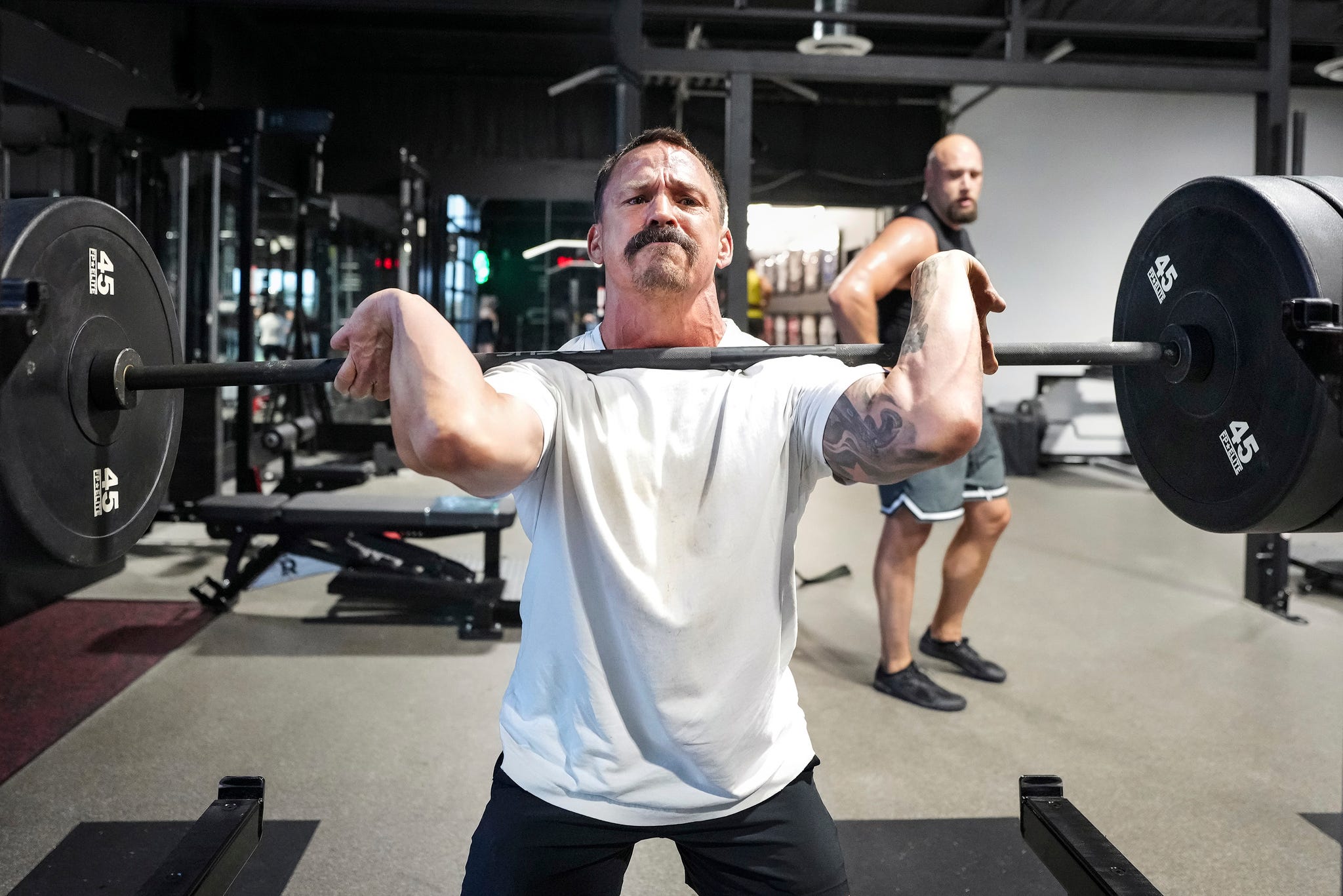 A man lifting a barbell in a gym, with another man in the background watching the exercise. A man lifting a barbell in a gym, with another man in the background watching the exercise.