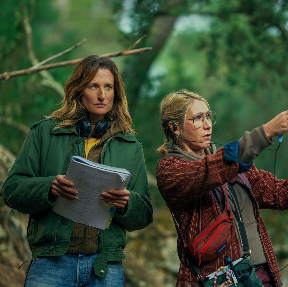 two women with scripts in a forest setting, discussing directions.
