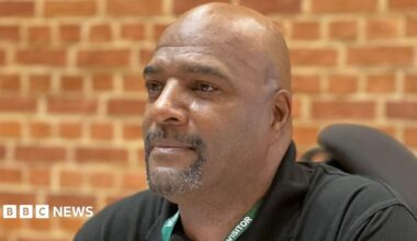 A man with short facial hair, wearing a black shirt and a green lanyard reading "visitor". He is pictured from the shoulders up and there is a brick wall behind him.