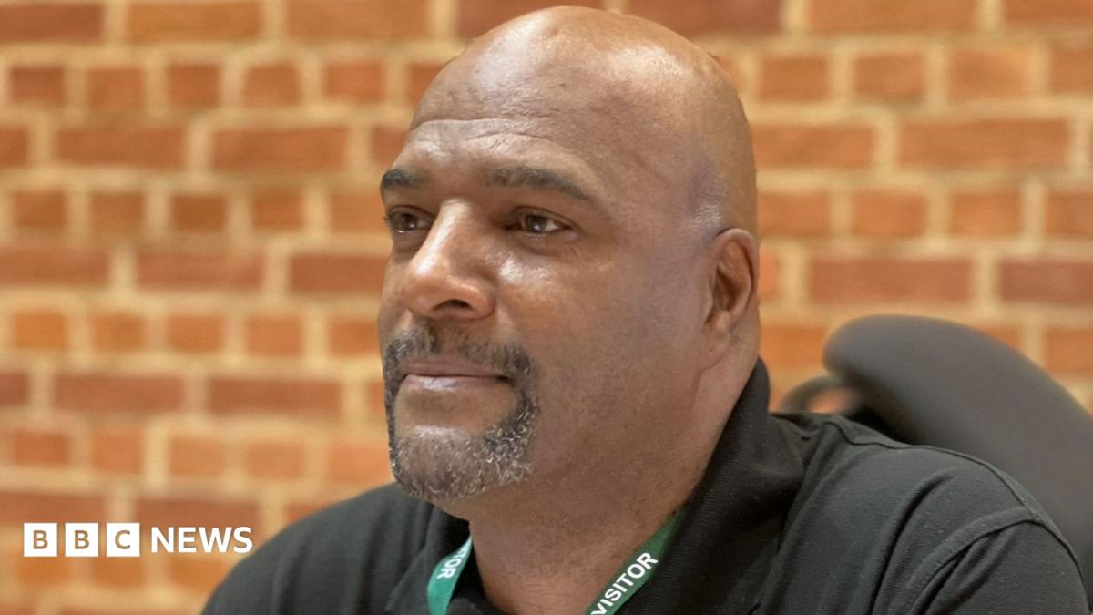 A man with short facial hair, wearing a black shirt and a green lanyard reading "visitor". He is pictured from the shoulders up and there is a brick wall behind him.
