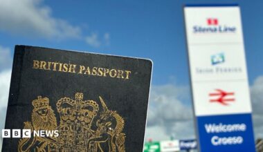 A dark blue British passport in the foreground with a sign welcoming people to Holyhead port, slightly out of focus in the background