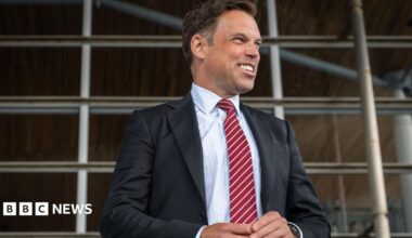 The image shows Ken Skates standing outside the Senedd. He is smiling and looking to his left. He is wearing a dark suit, white shirt and stripey red and white tie.