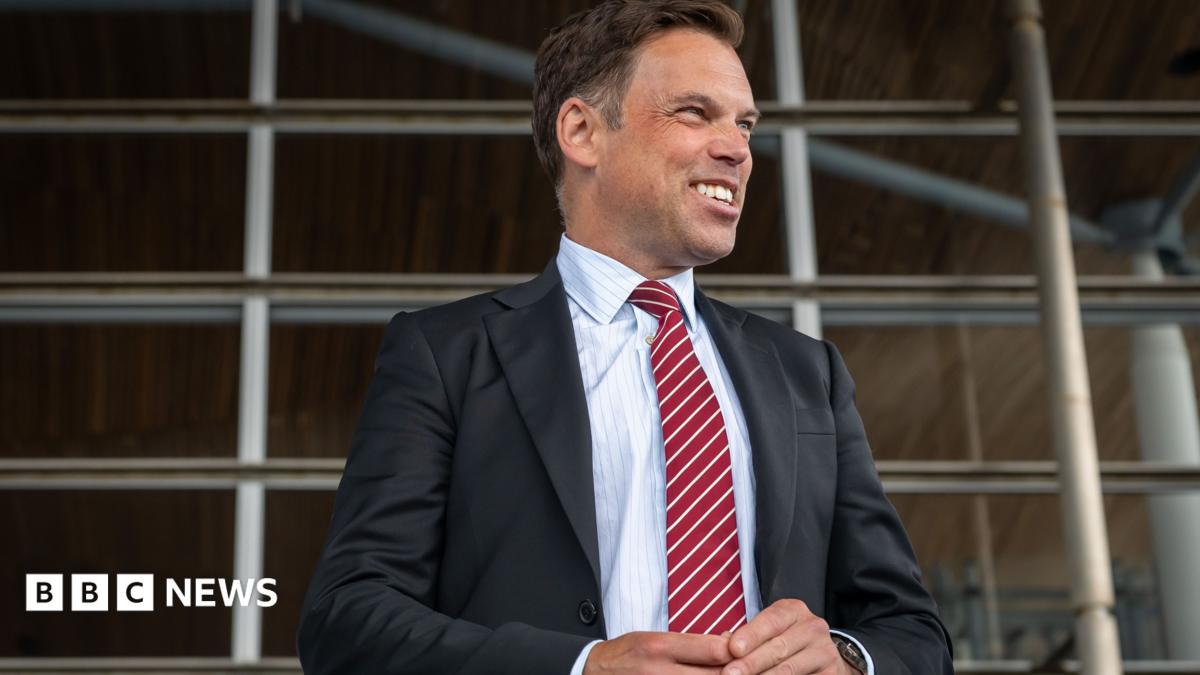 The image shows Ken Skates standing outside the Senedd. He is smiling and looking to his left. He is wearing a dark suit, white shirt and stripey red and white tie.