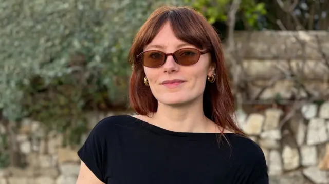 Alex Campbell is standing in front of a stone wall. She has long dark red hair and is wearing sunglasses and a black short-sleeved top.