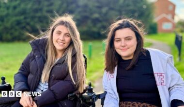 Two women in wheelchairs smile at the camera. There is a path and an area of green grass behind them. The woman on the left has long blond hair and is wearing a pale blue cardigan with a black puffa coat over the top. The woman on the right has shoulder length dark brown hair and is wearing a black top and an open, grey zip-up hoodie.