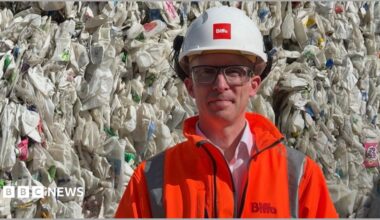 A man wearing safety glasses, an orange fluorescent jacket and white hard hat is standing in front of a huge stack of used plastic cartons