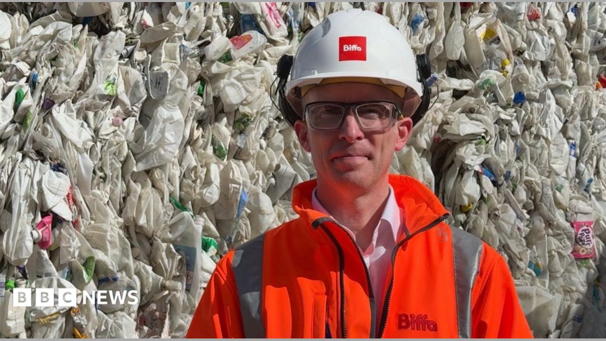 A man wearing safety glasses, an orange fluorescent jacket and white hard hat is standing in front of a huge stack of used plastic cartons
