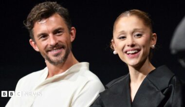 Jonathan Bailey, wearing a white open-necked shirt, and Ariana Grande in a black top and pearl stud earrings, at a Q&A on the film Wicked: For Good in Los Angeles. They are both laughing.