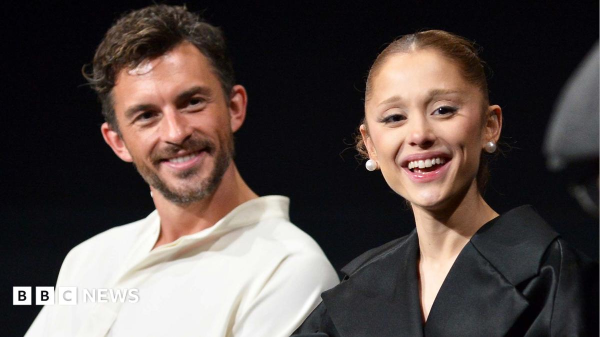 Jonathan Bailey, wearing a white open-necked shirt, and Ariana Grande in a black top and pearl stud earrings, at a Q&A on the film Wicked: For Good in Los Angeles. They are both laughing.