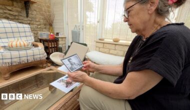 A woman in a black top and beige trousers is looking at a photo of her husband. The photo is black and white and he's in uniform. The woman, Anne, is sitting in a conservatory. There's soft furnishings such as a chair and foot stool with a coffee table directly in front of her. The back of the conservatory is lined with white open bricks along with the lower third next to the windows which have translucent blinds in front of them.