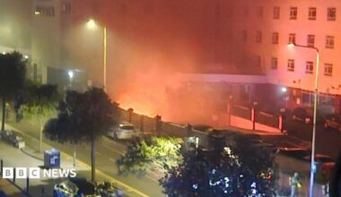A fire in the basement of a hotel. Flames can be seen from below the building, and emergency services can be seen in the foreground.