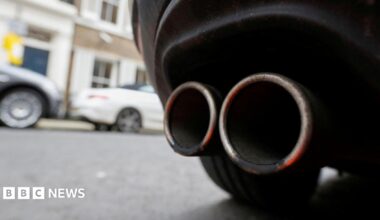 An exhaust pipe on the back of a car. A wheel can be seen, with two cars out of focus in the background