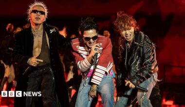 Three performers stand at the front of a stage holding handheld microphones during a live performance. One performer wears a long black coat and sunglasses, another wears a red military-style jacket with white trim and jeans, and a third wears a black studded leather jacket and ripped jeans. The stage is lit in red, with flames or flame-effect visuals displayed behind the performers.