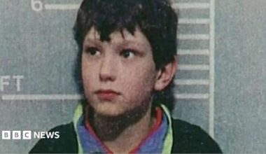 A police custody shot of a young Jon Venables, wearing a dark jumper, after his arrest. He is standing in front of a height chart showing he stands just over 4ft 6in tall.