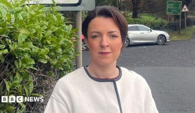 Liz Kimmins. She has dark brown hair tied back, wearing a white jacket with black lining on the edges. She is standing at a road junction with a car and road sign in the background, beside her is a bush.