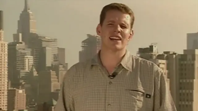A younger Rhun ap Iorwerth stands in front of the New York skyline in a short sleeved chequered shirt delivering a news package