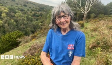 A woman is wearing glasses and a blue t-shirt, smiling at the camera while standing on a grassy hill.