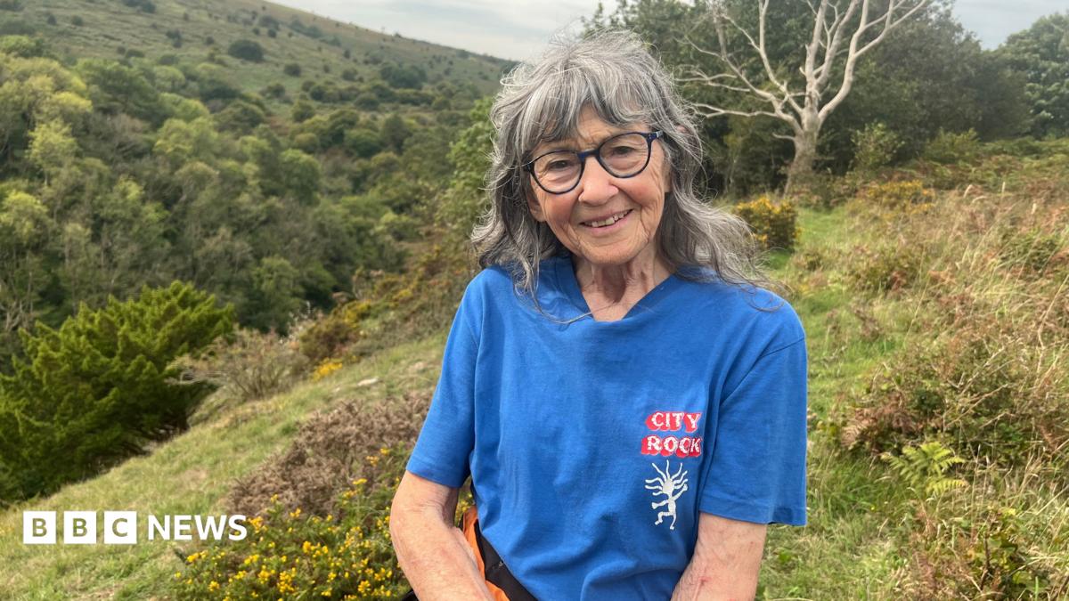 A woman is wearing glasses and a blue t-shirt, smiling at the camera while standing on a grassy hill.