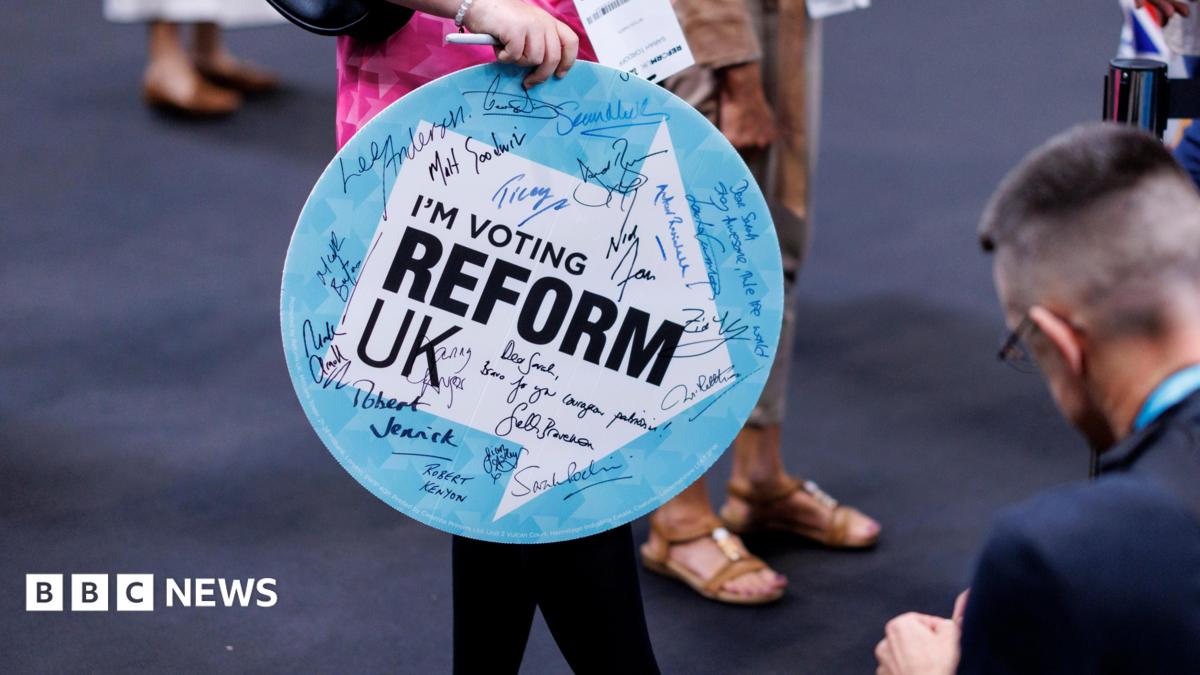 A woman holding a sign saying I'm Voting Reform UK enters the Reform conference. The sign has autographs on it from Reform politicians.