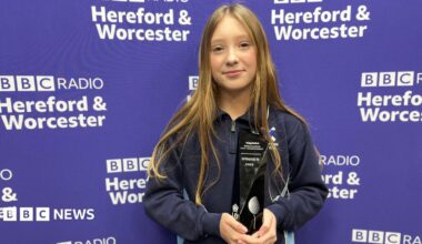 A 13-year-old girl with long dark blonde hair, wearing a dark blue quarter-zip jumper and holding a black trophy reading "winner 2025". She is standing in front of a purple backdrop reading "BBC Radio Hereford & Worcester"