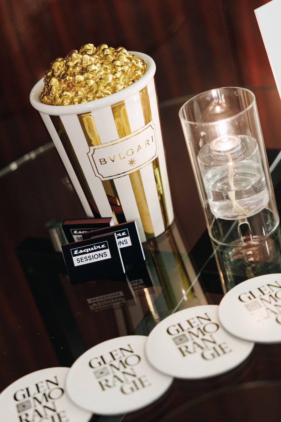 gold popcorn bucket labeled bvlgari, candle, coasters labeled glenmorangie, and esquire sessions matchboxes on a table.