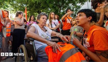 An elderly woman in a wheelchair is surrounded by protestors wearing orange in Madrid.
