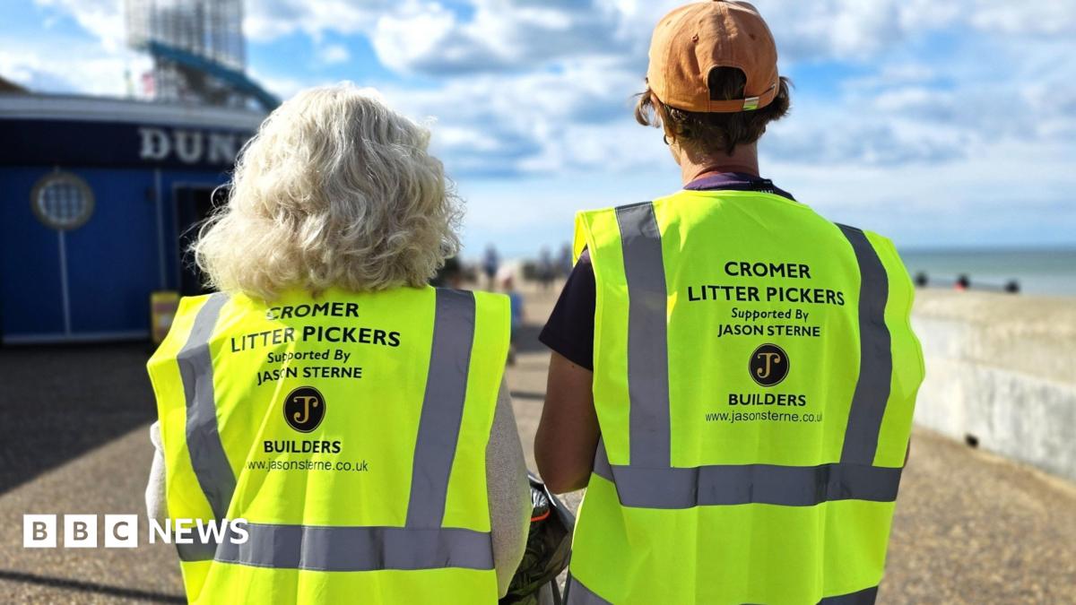 Two female volunteers wearing high-visibility “Cromer Litter Pickers” vests walk along Cromer seafront, carrying litter-collection equipment beside the beach. A sponsor's logo is displayed on the back of each vest, with the sea and promenade visible in the background.