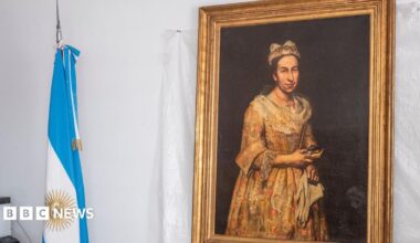 A portrait hangs on the wall with a gold frame on a white backdrop, next to it is an Argentinian flag rolled up