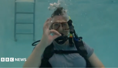 Luke Rogers is seen submerged in a pool, wearing a scuba mask and diving equipment. He is staring at the camera and making an expression with his fingers and hand to indicate things are going well
