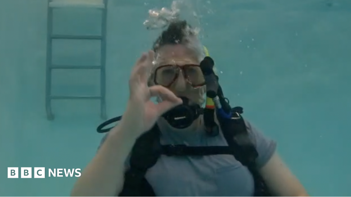 Luke Rogers is seen submerged in a pool, wearing a scuba mask and diving equipment. He is staring at the camera and making an expression with his fingers and hand to indicate things are going well