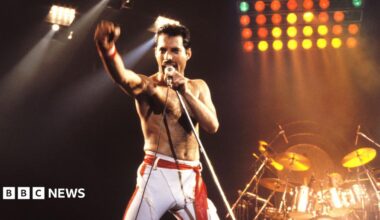 Freddie Mercury is pictured on a stage singing, with a microphone to his mouth and his arm raised. He is wearing white trousers. Behind him is a drum kit and lights.