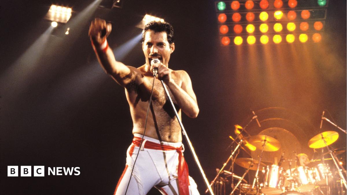 Freddie Mercury is pictured on a stage singing, with a microphone to his mouth and his arm raised. He is wearing white trousers. Behind him is a drum kit and lights.