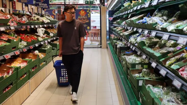 A man pulling a blue trolley basket through a supermarket fruit and veg isle