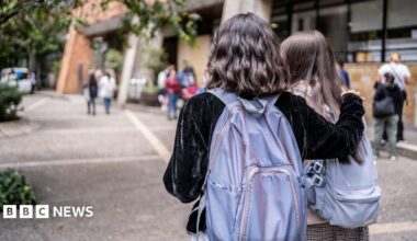 Two university students with purple backpacks walk across a university campus. Only the backs of their heads are visible. One has short wavy dark hair and wears a black velvet jacket. She has her right arm around the second student, who has long dark hair.