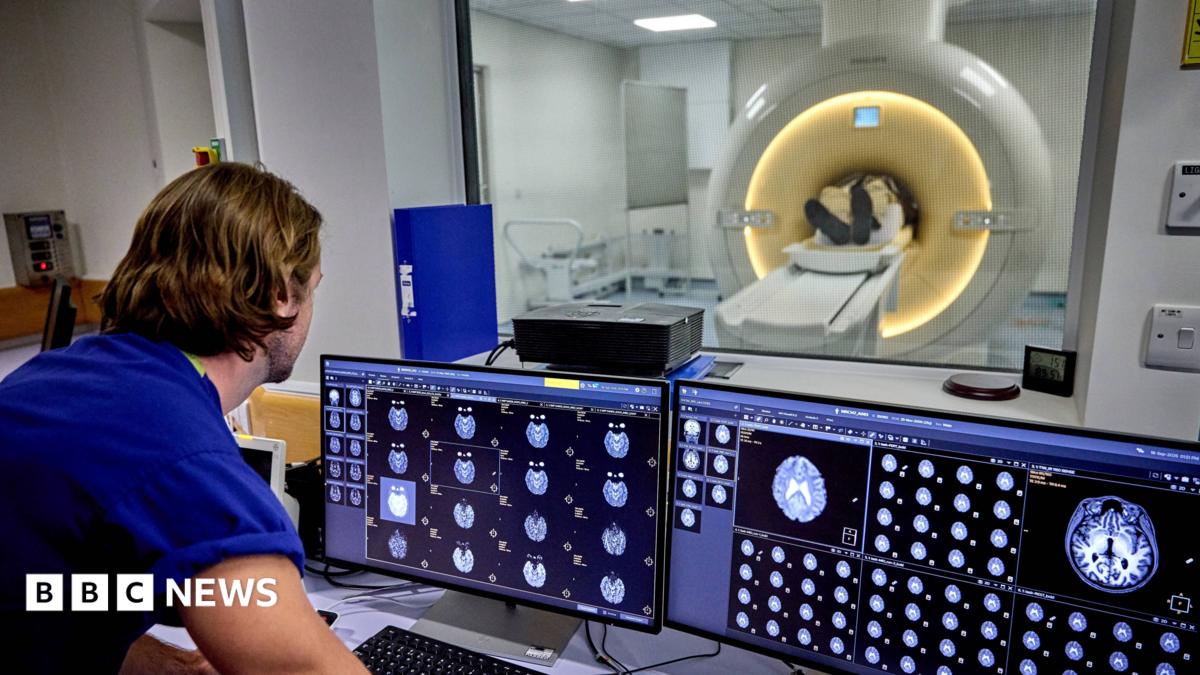 A man stands in front of brain scans and a MRI scanner through the glass window