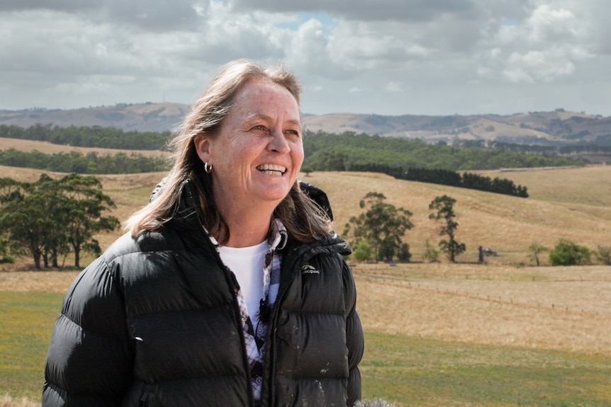 A woman wearing a black puffer jacket, looking to the side, smiling, with a rural farmland, trees and hills in the background