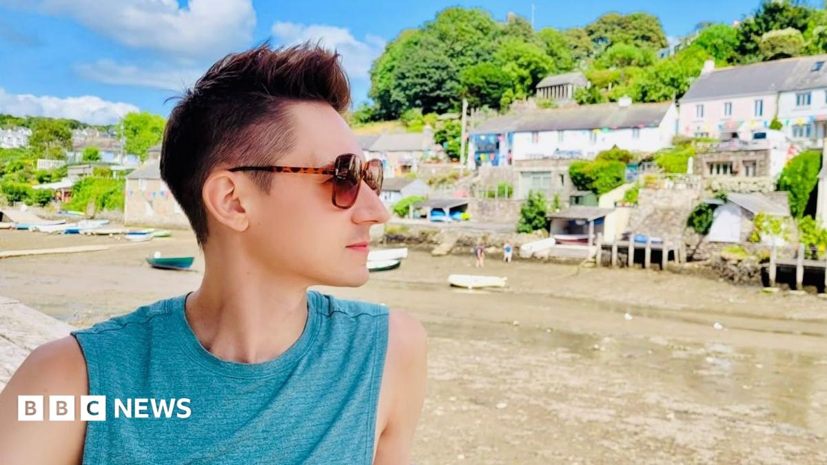 James Downs has short brown hair that is spiky on top. He is wearing a blue vest and brown sunglasses. He is looking out onto a river surrounded by boats and houses.