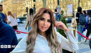 Sarah Khalifa wearing a white long-shirted with her folded left arm, the hand touching her long hair, sits by the side of a road inside a red-roped cordoned area, beyong which people walk on a road the other side of which has rows of sunbathed buildings