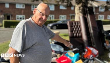 A man with a shaved head wears rectangular glasses and a grey t-shirt as he points to a large pile of fly-tipped rubbish under a tree on a green patch of land with terraced houses in the background. Among the items are a leather-effect bedstead and bags full of rubbish strewn on the ground.