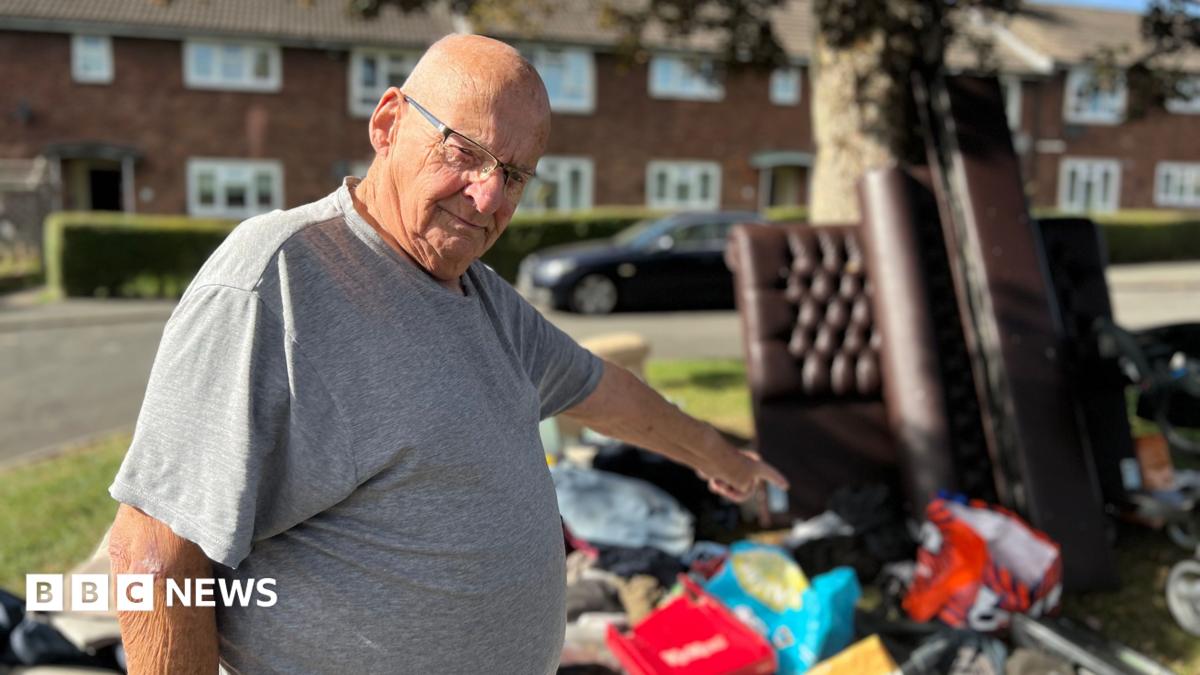 A man with a shaved head wears rectangular glasses and a grey t-shirt as he points to a large pile of fly-tipped rubbish under a tree on a green patch of land with terraced houses in the background. Among the items are a leather-effect bedstead and bags full of rubbish strewn on the ground.