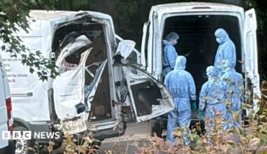 people in blue hazmat suits stand behind a white van next to another white van with the back doors blown off
