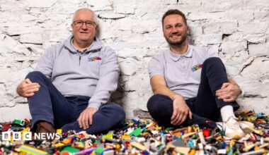 Two men wearing WeBuyBricks tops sit on top of a large pile of multicoloured Lego pieces against a white brick wall.
