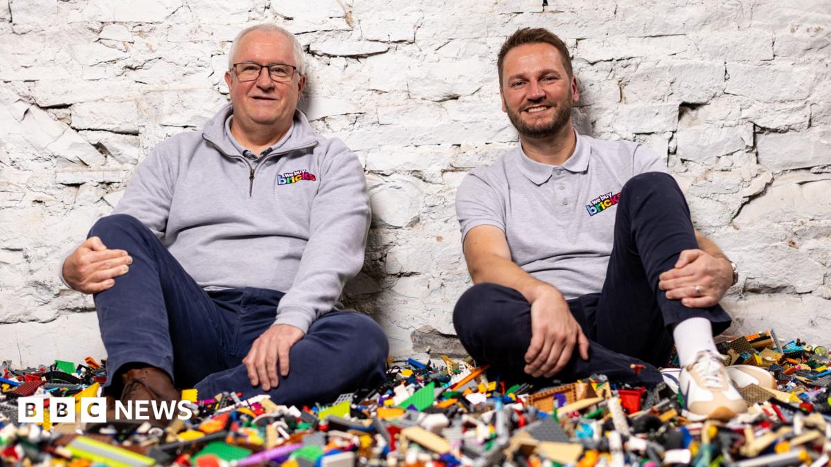 Two men wearing WeBuyBricks tops sit on top of a large pile of multicoloured Lego pieces against a white brick wall.