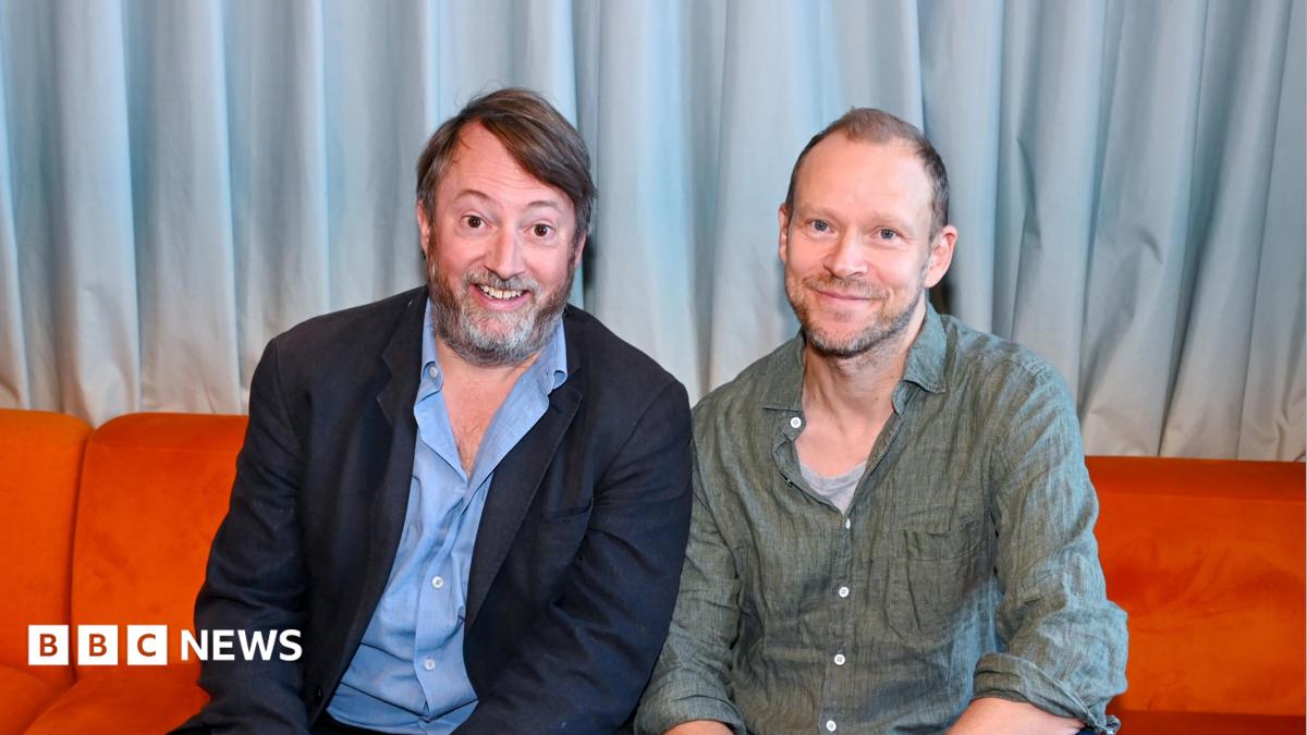 David Mitchell (left) and Robert Webb sitting on a red sofa and smiling directly into the camera