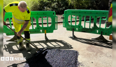 A man wearing high-vis uniform is using a tool to cover a pothole. There are green barriers around him and bushes can be seen in the background.