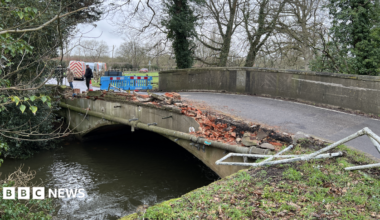 A general view picture of the bridge, which has seen its side closest to the camera totally flattened, with metal railings bent out of place and bricks scattered nearby.