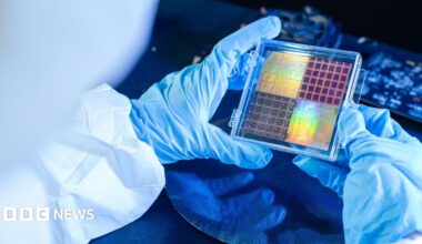 An engineer in a cleanroom, wearing blue gloves and white overalls, inspects a sheet of semiconductors